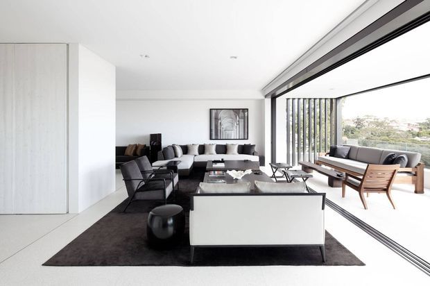 Bright, open-plan contemporary living room with a restrained monochrome palette. The space features a large dark area rug, low-profile sofas and armchairs, a central coffee table, a white-backed console or sofa element, and extensive floor-to-ceiling glazing with slim dark frames. A furnished glazed terrace or balcony is visible beyond the windows, overlooking a suburban landscape.