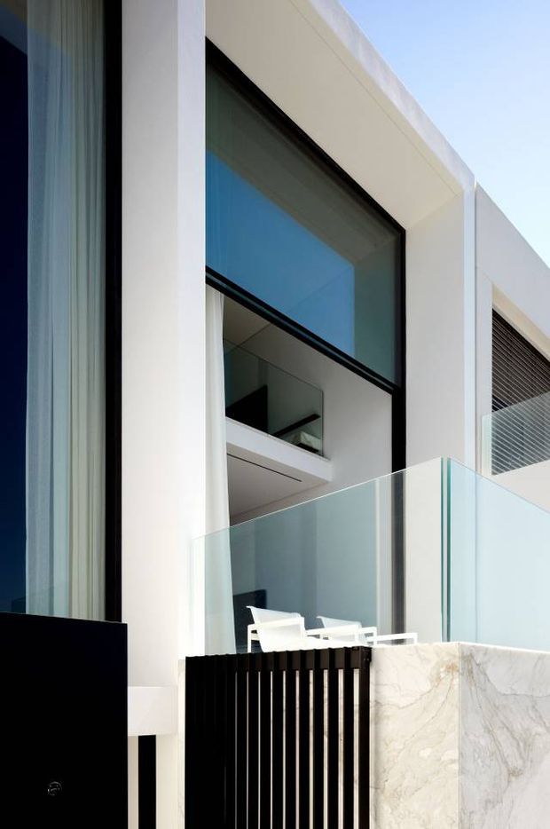 Close-up exterior view of a contemporary multi-level building with a crisp white facade, large black-framed glazed opening, transparent glass balcony balustrade, black vertical privacy screen, and pale veined stone wall. White outdoor seating is visible behind the balcony glazing. The composition emphasizes strong geometric lines, full-height glazing, and refined modern materials.