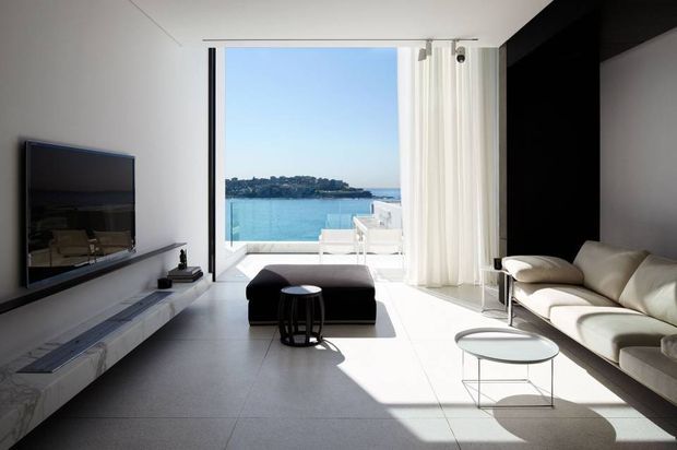 Bright minimalist living room with floor-to-ceiling glazing opening to a waterfront view. The room features a wall-mounted television, long floating stone or concrete-look media console, cream sectional sofa, black ottoman, round glass coffee table, large-format pale flooring, and sheer white curtains. Strong sunlight creates a clean, high-contrast interior with a contemporary coastal atmosphere.