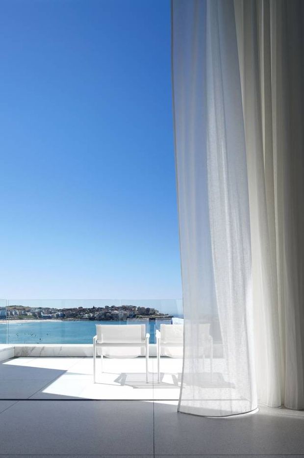 Bright, minimalist interior opening onto a waterfront balcony or terrace. Floor-to-ceiling sheer white curtains frame full-height glazing, with a pair of white outdoor chairs on a glass-balustraded terrace overlooking clear blue water and a built-up shoreline. The space has pale large-format flooring, strong sunlight, and an airy coastal atmosphere.