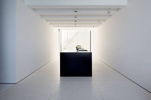 Minimalist monochromatic interior corridor or gallery-like space with white walls, a white exposed-beam ceiling, pale tiled flooring, and a centrally placed black rectangular island or plinth. A small dark fixture is visible at the far end, with bright glazing or an opening beyond.