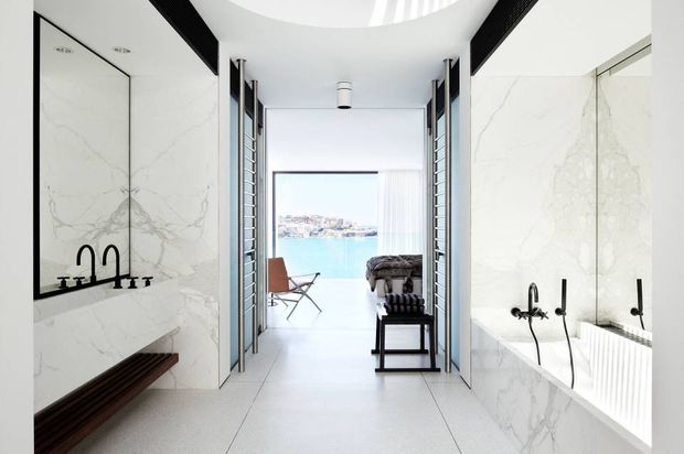 Bright, luxurious contemporary bathroom corridor with white marble-effect wall surfaces, a freestanding or built-in bathtub with black fixtures, and a double vanity with black taps and large mirrors. Full-height glazed or louvred openings lead toward a waterfront view and seating area beyond. The space has a clean linear layout, pale tiled flooring, dark timber bench seating, and strong black-and-white contrast.