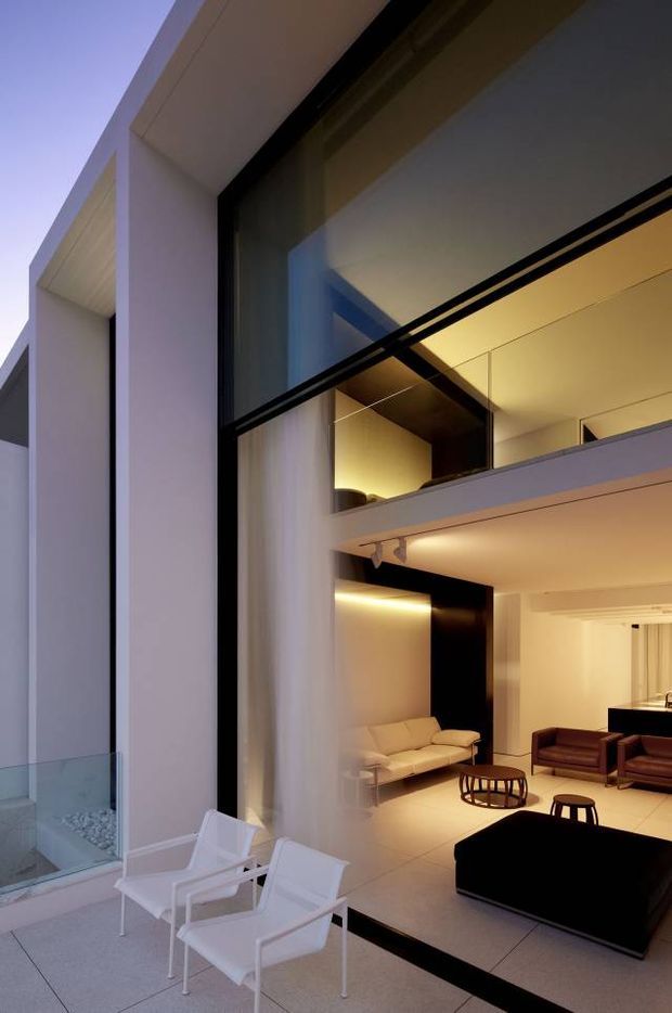 A contemporary multi-level residence viewed from an outdoor terrace through a large double-height glazed opening. The composition shows a minimalist white facade, expansive dark-framed glazing, an upper-level mezzanine or landing, and a warm modern living room with sofas, armchairs, a coffee table, and concealed architectural lighting. White outdoor lounge chairs sit on the terrace in the foreground.