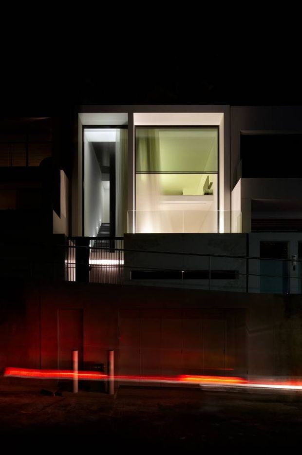 A dramatic nighttime exterior photograph of a contemporary multi-level residence. The facade features large illuminated windows, strong rectangular volumes, dark wall surfaces, and a glass-fronted balcony or terrace with metal railings. Long-exposure vehicle light trails create vivid red and white horizontal streaks across the street in the foreground.