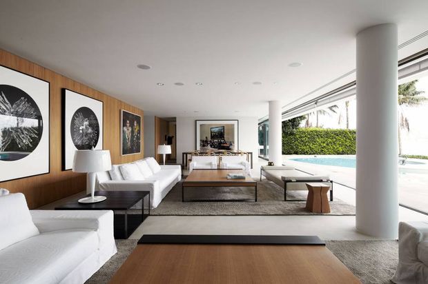 A bright, spacious contemporary living room with white sofas and armchairs arranged around low dark coffee tables, set against a warm timber-panelled feature wall with large monochrome artworks. Full-height glazing opens the room toward a pool terrace, while white cylindrical columns, recessed ceiling lights, and a neutral textured rug reinforce the clean modern design.