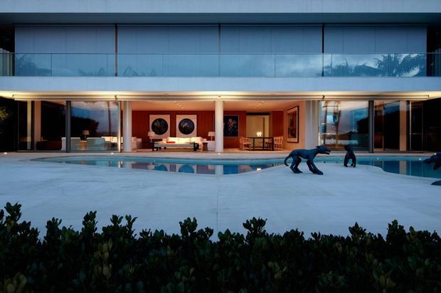 Wide exterior view of a sophisticated two-storey modern residence at dusk, with extensive floor-to-ceiling glazing, a long projecting upper balcony, slender white columns, and a covered ground-level terrace opening onto a broad swimming pool and pale paved pool deck. Warm interior lighting contrasts with the cool blue exterior light; dark landscaping frames the foreground.