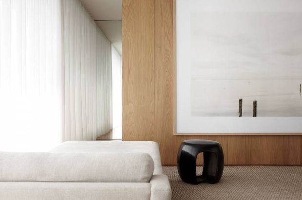 A serene contemporary living room featuring a low cream upholstered sofa, full-height sheer curtains, warm natural timber wall panelling and built-in joinery, a large minimalist framed artwork, and a sculptural black side stool. The room has a restrained neutral palette, soft natural light, and a calm, refined atmosphere.