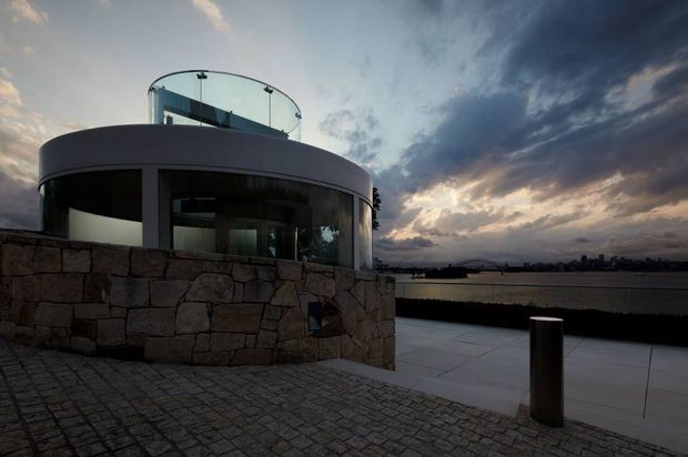 Exterior dusk view of a contemporary rounded pavilion or house beside a broad waterfront or open landscape. The building has curved white rendered walls, extensive dark glazing, a transparent curved glass balustrade at roof level, and a substantial irregular natural-stone retaining wall. A paved terrace and cobbled foreground lead toward the water, with dramatic cloud-filled evening sky.