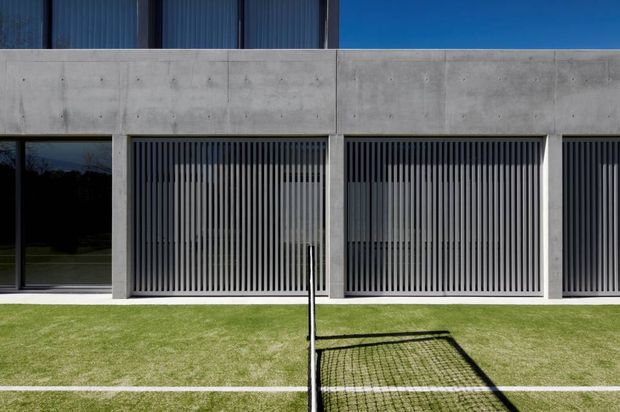 A minimalist contemporary building facade viewed from an outdoor tennis court. The lower level is formed in smooth raw concrete with large glazed openings screened by closely spaced vertical metal fins, while the upper level has dark vertically ribbed cladding and expansive dark glazing. A neatly maintained grass court occupies the foreground, with a tennis net casting a strong shadow.