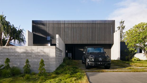 Contemporary two-storey house exterior with a dark vertically ribbed upper facade cantilevered over a recessed entry and garage. The building combines concrete block boundary walls, smooth pale rendered surfaces, dark glazing, and layered landscaping with a broad driveway in the foreground.