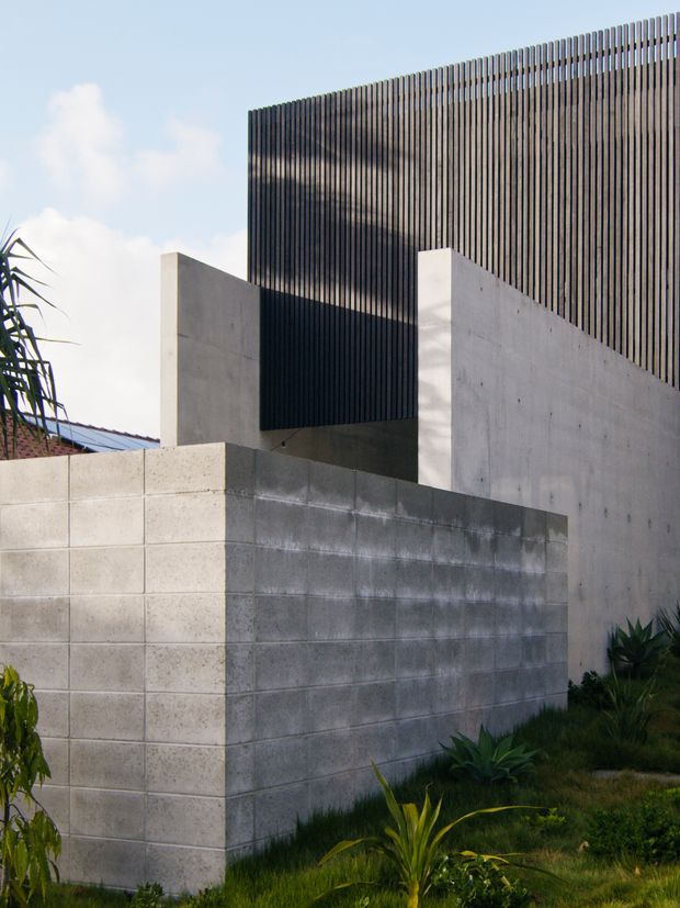 Exterior architectural photograph of a modern building enclosed by pale concrete block and smooth concrete walls, with a tall dark vertical slatted screen forming the upper facade. The composition includes minimalist geometric forms, landscaped planting, and a clear sky.