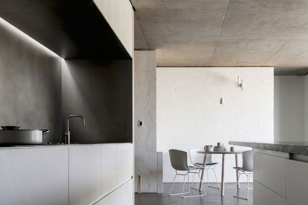 A minimalist open-plan kitchen and dining area with white flat-panel cabinetry, a dark grey splashback and benchtop, integrated under-cabinet lighting, and a concrete ceiling. A small round dining table with modern chairs sits beyond the kitchen, with exposed white-painted brickwork and polished grey flooring contributing to the restrained industrial-modern aesthetic.