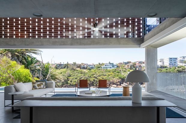 A bright, open-plan living room with a large sectional sofa facing expansive glazing and a scenic leafy suburban outlook. The room features a dark built-in console or table, sculptural dome-shaped lamp, concrete ceiling and structural beams, and a perforated brick privacy screen above the glazing. The restrained furnishings and strong indoor-outdoor connection create a refined contemporary atmosphere.
