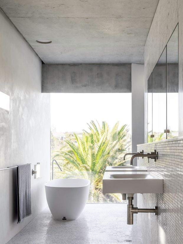 A bright, minimalist bathroom with a freestanding white bathtub positioned beside a large opening overlooking palm foliage. Two wall-mounted white rectangular basins with exposed metal plumbing sit beneath a full-width mirrored wall. The room has a raw concrete ceiling and upper wall beam, pale textured plaster walls, and small light-grey mosaic floor and wall tiles. Black or dark metal tapware, towel rail, and fixtures provide subtle contrast.