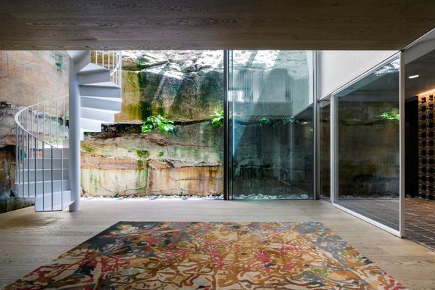 A contemporary interior living space with a full-height glazed wall opening onto a rugged rock and planted courtyard. A sculptural white spiral staircase rises at the left, while a dark timber ceiling, pale timber flooring, expansive glass panels, and a large abstract patterned rug define the room. Reflections in the glazing extend the view of the interior and courtyard.