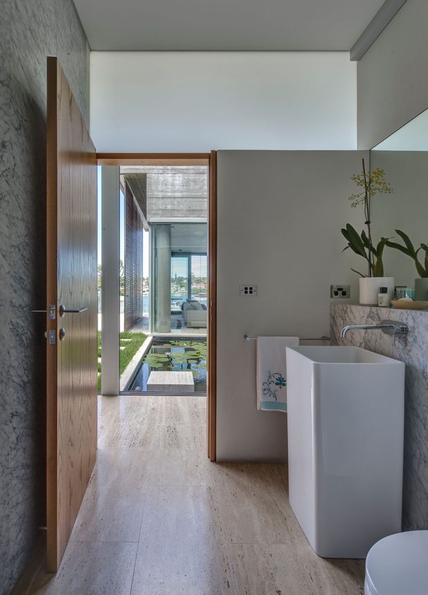 A refined contemporary bathroom viewed toward an open timber door and an internal courtyard. The room features a freestanding white rectangular basin, wall-mounted chrome tapware, a marble-patterned stone vanity or shelf, pale natural-stone flooring, a textured dark stone wall, timber door and trim, and a large mirror. A water feature with lily pads and additional modern glazing is visible beyond the doorway.