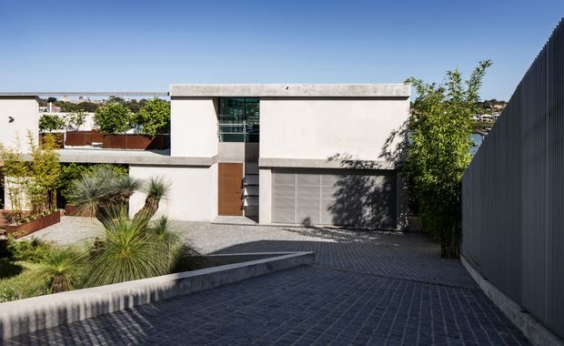 Exterior view of a minimalist modern residence, showing a cubic rendered-concrete facade, narrow horizontal roofline, large upper-level glazing, and a broad grey garage door. A sloping dark-paved driveway leads toward the house, bordered by low concrete walls and drought-tolerant landscaping. A tall dark vertical fence frames the right side, with neighbouring homes visible in the background.