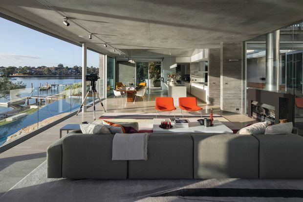A spacious contemporary open-plan living room overlooking the water, with full-height glazing, a concrete ceiling and floor, a sleek kitchen, fireplace, dining area, and large grey sectional sofa. Bright natural light and vivid orange accent chairs contrast with the restrained concrete and neutral palette.