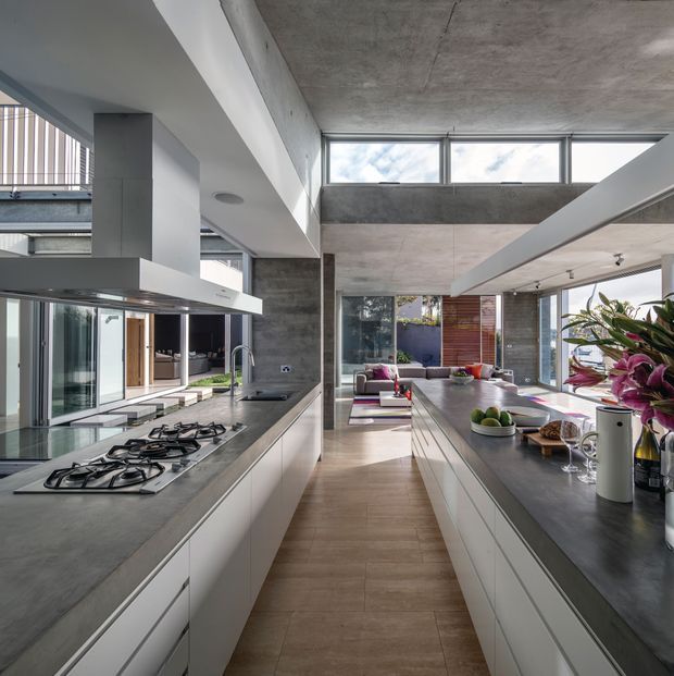Contemporary open-plan kitchen in a spacious, light-filled home. The room features long parallel runs of white flat-panel cabinetry, dark grey concrete-look benchtops, an integrated gas cooktop and rangehood, large glazed openings, clerestory windows, exposed concrete ceilings and columns, and timber-toned flooring. A living area is visible beyond the kitchen, with abundant daylight and greenery outside.
