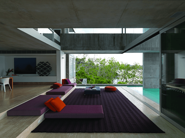 A dramatic contemporary split-level living room with exposed concrete ceilings and walls, expansive glazing, and a strong indoor-outdoor connection to a terrace and greenery beyond. The room features low modular purple seating arranged around a dark textured rug and coffee table, with orange cushions and an orange ottoman adding vivid accents. A dining/work area is visible to the left, while a turquoise-toned pool or water feature appears through the glazing on the right.