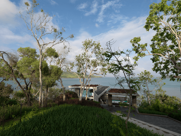 A low-slung modern pavilion-style building set into a lush, landscaped coastal hillside, with expansive views toward calm blue water. The structure has exposed concrete elements, dark roof planes, broad open glazing, and an angular sculptural form, partially screened by mature trees and dense planting. A paved path and terrace lead through the foreground garden under a bright, lightly clouded sky.