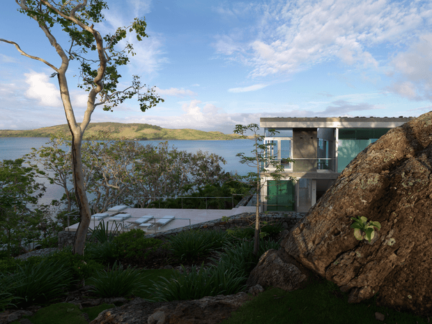 A modern multi-level home set into a steep, rocky waterfront site, viewed from landscaped grounds. The building has a flat roof, large glazed openings, concrete-toned surfaces and projecting terraces overlooking the blue water and distant hills. Native-style planting, exposed rock and a pool or terrace area reinforce the integration with the natural landscape.