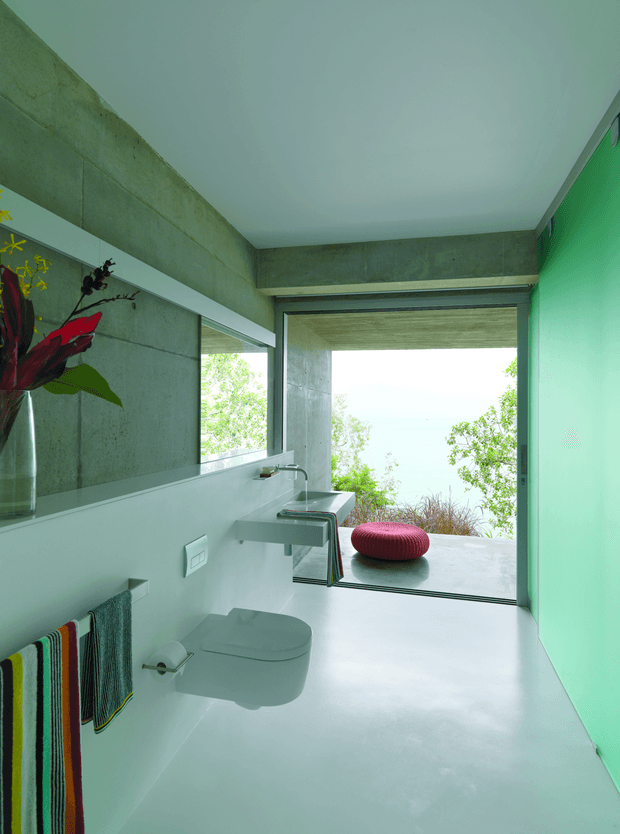 Contemporary minimalist bathroom with a wall-mounted white toilet, integrated wall-mounted basin and chrome tap, smooth pale flooring, exposed grey concrete wall and ceiling beams, and a vivid green accent wall. A large frameless opening connects visually to a planted outdoor terrace, with a red upholstered ottoman visible outside. A wall mirror reflects the basin area and a colourful towel adds a small accent.