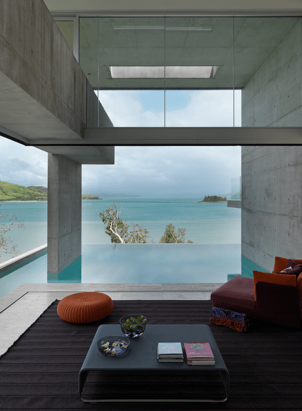 A dramatic modern living room opening directly onto a turquoise waterfront, framed by exposed raw concrete walls, columns, and ceiling. Full-height glazing and a large overhead skylight create strong indoor-outdoor connection and expansive water views. The room contains a low dark coffee table, black textured rug, deep burgundy sectional seating, and a sculptural orange round ottoman. A turquoise pool or water feature appears beyond the room, with distant green hills and small islands visible outside.