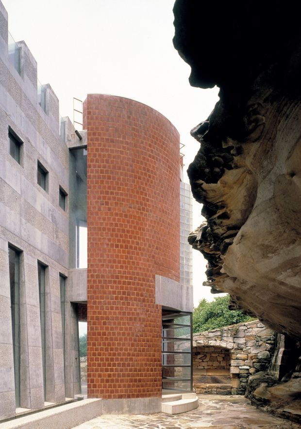 An exterior architectural view showing a sculptural red brick cylindrical tower or curved wall set between pale textured masonry/concrete buildings and a dramatic natural rock overhang. Glazed vertical sections, narrow windows, exposed metal railings, stone retaining walls, and irregular stone paving create a strong contrast between contemporary construction and the rugged rock formation.