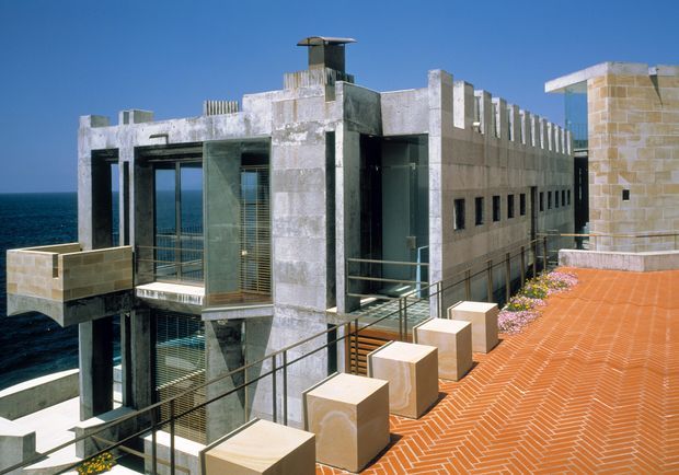 Exterior view of a striking multi-level coastal building beside the sea. The structure features raw concrete volumes, extensive glazing, narrow horizontal windows, strong vertical fins, rooftop elements, and a large terracotta herringbone-paved terrace with pale stone-clad walls and balustrade blocks.