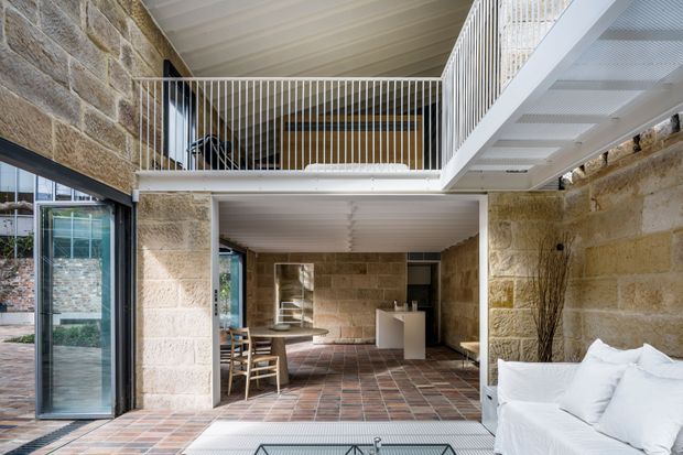 Double-height interior of a contemporary stone-clad residence, showing a mezzanine landing with slim white metal balustrades, exposed sandstone walls, white-lined ceilings, expansive glazing and a tiled floor. The lower level combines a living area in the foreground with a dining and kitchen zone beyond; a white sofa is visible at right and an open stair/mezzanine structure creates strong vertical volume.