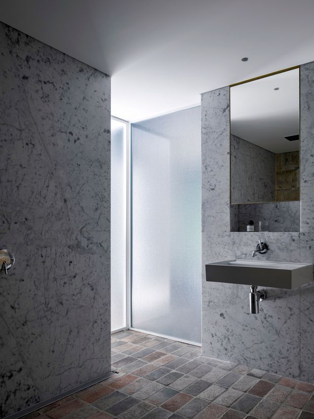 Contemporary bathroom with grey veined stone wall surfaces, a wall-mounted rectangular basin, large frameless mirror, exposed chrome plumbing, and a translucent glass shower enclosure. The floor uses small square tiles in muted grey, brown, and mauve tones, while recessed ceiling lights and a narrow brass-toned mirror trim add subtle detailing.