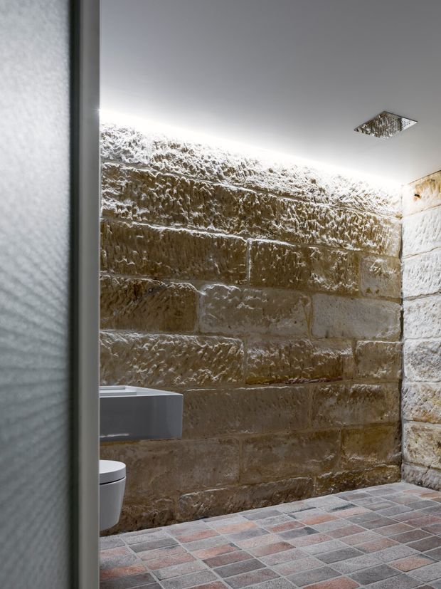 A compact bathroom viewed through a doorway, featuring rough textured natural stone block walls, a wall-mounted toilet partially visible at left, a square ceiling showerhead, and a small-format multicoloured tiled floor. Bright concealed perimeter lighting highlights the stone texture, creating a rustic yet refined atmosphere.