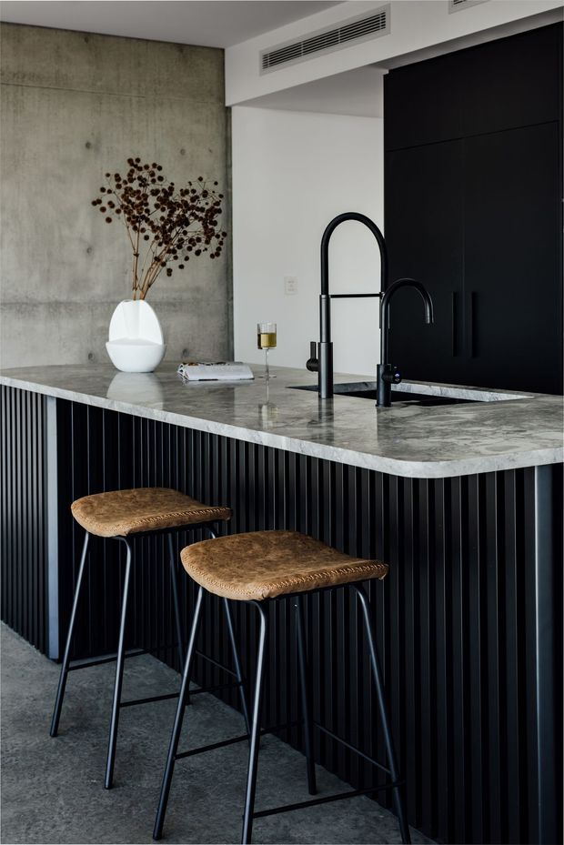 A sophisticated contemporary kitchen featuring a large grey-veined stone island with an integrated sink and two matte-black gooseneck faucets. The island base is clad in vertically fluted black cabinetry, paired with tan woven leather bar stools and dark cabinetry. A raw concrete feature wall, minimalist decor, and concealed linear air-conditioning vents complete the refined industrial-inspired interior.