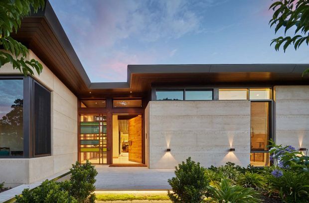 A contemporary single-storey residence viewed from the landscaped front approach at dusk. The facade combines smooth pale concrete walls, dark metal-framed glazing, warm timber soffits and timber entry detailing beneath broad flat roof planes. Recessed exterior lighting, planted garden beds and a sheltered recessed entry create a refined, welcoming composition.