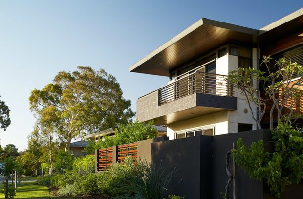 Exterior view of a contemporary multi-storey residence with projecting flat rooflines, a deep timber-lined eave, concrete and dark cladding, and a small upper-level balcony enclosed by horizontal metal balustrades. Dark boundary fencing, timber screening, landscaped planting, and mature trees frame the house.