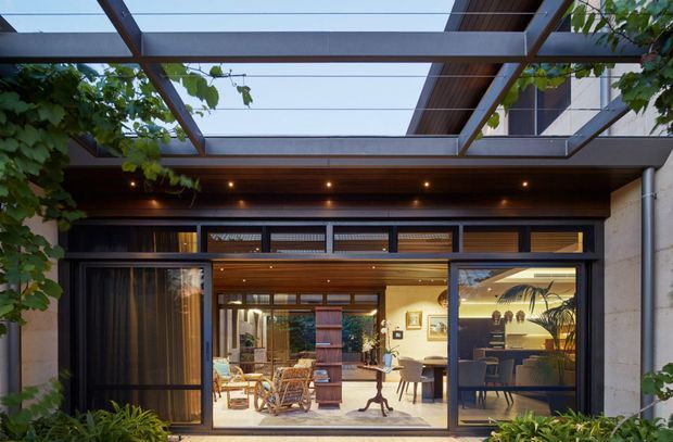 A contemporary courtyard-facing residential facade viewed from outdoors, with large black-framed glazed sliding doors opening onto a warmly lit interior living and dining area. A timber-lined soffit with recessed downlights sits beneath a translucent pergola roof, while climbing vines and lush planting soften the architectural composition. The interior shows timber joinery, lounge seating, a dining table and chairs, and a compact kitchen or bar area beyond.
