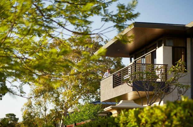 Exterior view of a contemporary multi-level residential home partially screened by mature landscaping. The building features a broad low-pitched roof overhang, dark-framed glazing, light masonry or rendered walls, and a prominent upper-level balcony with horizontal metal balustrading. Warm sunlight and abundant greenery give the scene a relaxed suburban character.