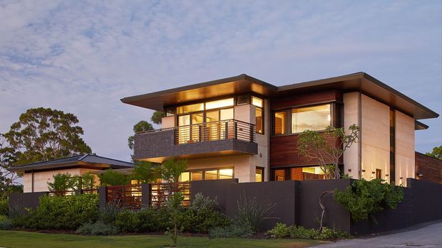 Twilight exterior view of a contemporary multi-level residence with broad low-pitched roof planes, warm illuminated glazing, rendered or concrete-toned wall panels, dark horizontal cladding, timber-toned feature walls, and a projecting upper-level balcony with metal balustrade. The home is screened by landscaped planting and dark boundary walls.