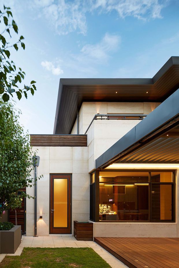 Exterior view of a sophisticated contemporary multi-level residence, featuring pale stone or concrete wall panels, dark metal roof and balcony elements, warm timber soffits, a timber deck, and extensive glazed openings. A compact landscaped courtyard and entry are visible, with warm interior lighting glowing through the large window.