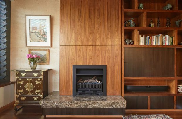 A richly detailed living room feature wall with floor-to-ceiling walnut-toned timber joinery, an integrated fireplace, open display shelving, bookshelves, and concealed media storage. A polished dark stone hearth extends across the base, contrasting with a textured beige wall and eclectic decorative furnishings. The composition is warm, refined, and layered, with strong mid-century influences.