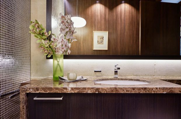 A refined contemporary bathroom vanity is shown close-up. The room features a floating dark timber-grain vanity with long horizontal metal handles, a polished brown marble benchtop with an integrated oval basin, chrome wall-mounted tapware, and a large mirror framed by dark timber cabinetry. A horizontal illuminated wall recess sits beneath the mirror, while small-format dark mosaic tiles clad the left wall. Decorative orchid flowers and a framed artwork add softness and detail.