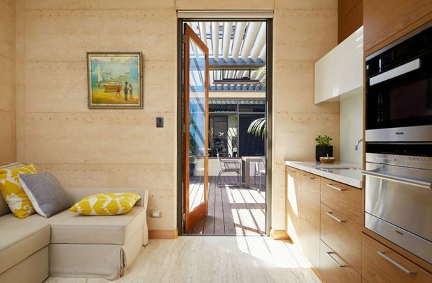 A bright, compact contemporary kitchen and adjoining living area. The kitchen features warm timber cabinetry, a white benchtop, integrated stainless-steel appliances, and white overhead joinery. A glazed timber-framed door opens to a sunlit outdoor courtyard or terrace. The adjacent living area includes a light sectional sofa with yellow patterned cushions, while the walls and floor have warm pale stone-like finishes.