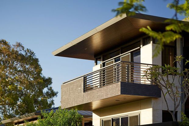 Exterior architectural photograph of a contemporary multi-level home, focused on a projecting upper-floor balcony with horizontal metal balustrades, light masonry walls, dark timber-lined soffits, and extensive glazing. The building has a broad low-pitched roof overhang and is surrounded by mature landscaping.