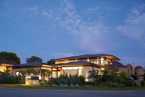 Twilight exterior photograph of a large contemporary multi-level residence with broad low-pitched metal roof planes, warm recessed lighting, extensive glazing, pale rendered walls, landscaped front garden, and a suburban street frontage. A neighbouring home is visible at the right edge.