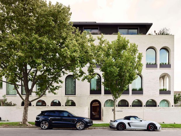 Street-facing view of a multi-storey contemporary building with a pale masonry facade, repeated arched windows and openings, dark upper-level cladding, and a projecting flat roof. Mature trees partially screen the facade, with a low perimeter wall and residential street in front.