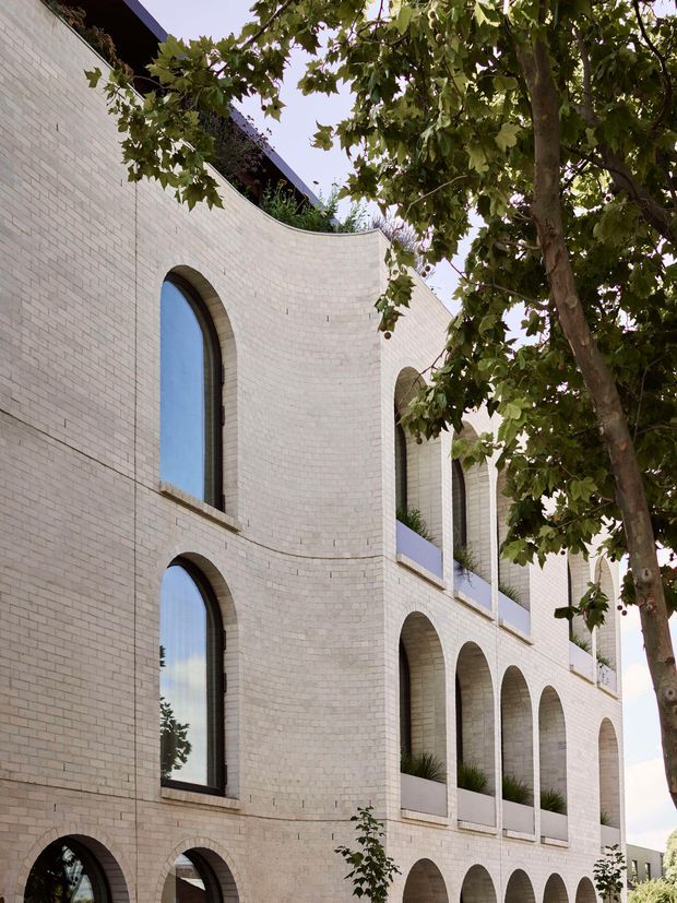 Exterior architectural photograph of a multi-storey building with a pale cream brick facade, strongly curved wall volumes, and repeated tall arched windows and recessed arcades. Dark-framed glazing, planted balcony recesses, and mature street trees create a refined contemporary facade with classical geometric references.