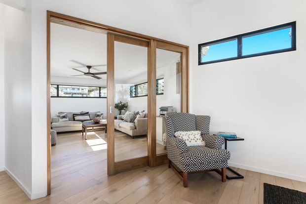 Bright contemporary interior showing a compact sitting or living area with a bold black-and-white patterned armchair, light timber flooring, white walls, and a large timber-framed mirrored sliding partition. The mirror reflects an adjacent living room with a sofa, coffee table, ceiling fan, and broad windows. High horizontal clerestory-style glazing provides natural light.