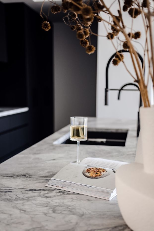 Close-up view of a refined contemporary kitchen featuring a veined light marble island benchtop, dark cabinetry, an undermount sink with a sculptural black gooseneck tap, and a white vase with dried botanicals. An open design book and wine glass add a styled, sophisticated atmosphere.