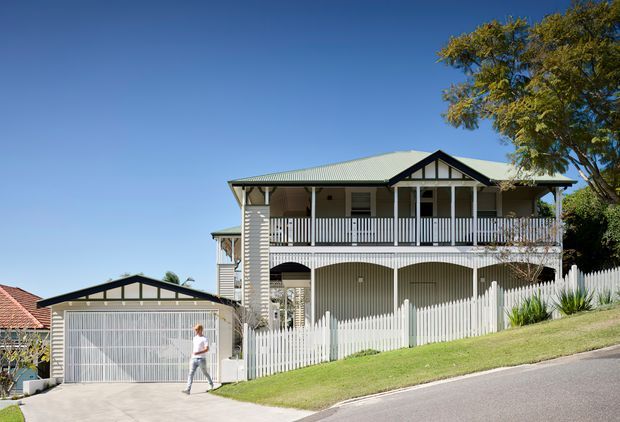 A two-storey suburban house on a sloping site, featuring a light-coloured corrugated metal roof, raised verandahs and balconies, white vertical picket fencing, decorative gable timber detailing, and a detached double garage. The facade combines rendered masonry, horizontal weatherboard-like cladding, and painted timber balustrades. A broad-leaf tree and landscaped front verge frame the property.